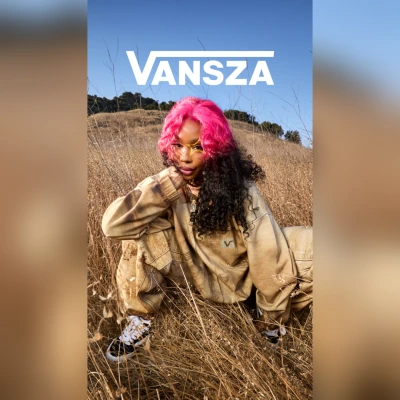 Model with pink hair in a tan sweatsuit sitting in a field of dry grass